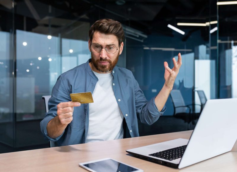 Cheated and disappointed businessman working inside office, man holding bank credit card and trying to make a purchase in online store, online fraud and deception Cheated and disappointed businessman working inside office, man holding bank credit card and trying to make a purchase in online store, online fraud and deception.